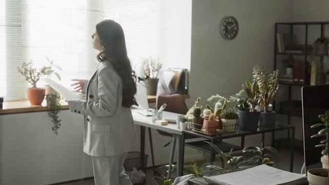 Hispanic young adult woman wearing headset pacing in office while holding documents and speaking. Clip is ideal for corporate communication, customer support training, or remote work marketing