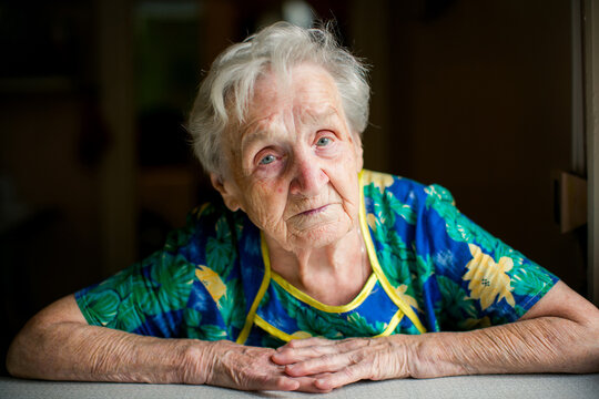 Portrait of an elderly woman resting her arms on a table and looking at the camera indoors in natural light