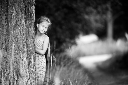 Black and white portrait of a young girl peeking from behind a tree and looking at the camera outdoors