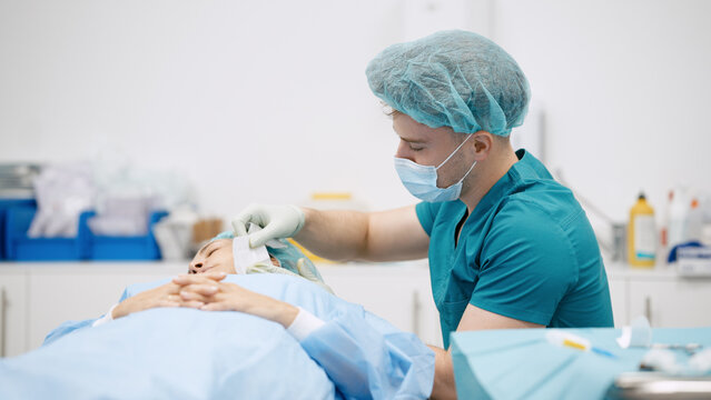 Surgeon wearing scrubs and mask preparing female patient's head for a medical procedure in a modern hospital operating room