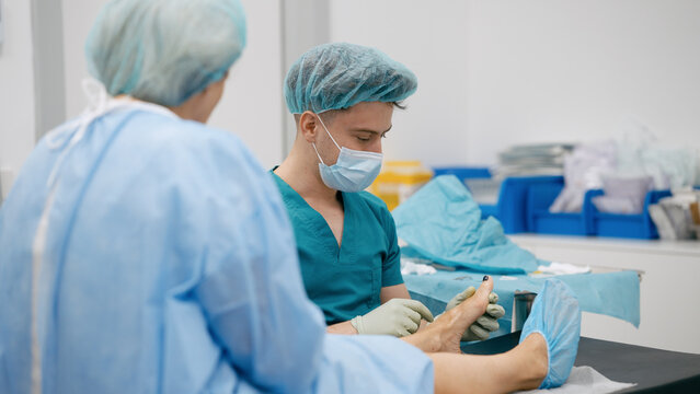Male doctor in scrubs and mask examining a patient's foot in a modern hospital setting, providing medical care