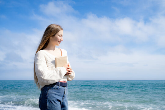 Young woman walking along the beach with a book on a sunny day, enjoying the sea and peaceful surroundings. Casual coastal lifestyle, reading and slow living concept.