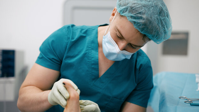 Medical professional in scrubs, mask, and cap examining or treating a patient's foot in a sterile hospital operating room