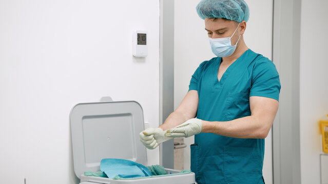 Surgeon wearing scrubs, hairnet, and face mask putting on sterile gloves, preparing for a medical procedure in a hospital