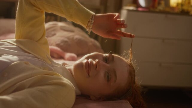 Portrait of Caucasian teenage girl lying on bed, twirling red hair and smiling at camera. Clip is perfect for youth mental health campaigns or lifestyle brand advertising