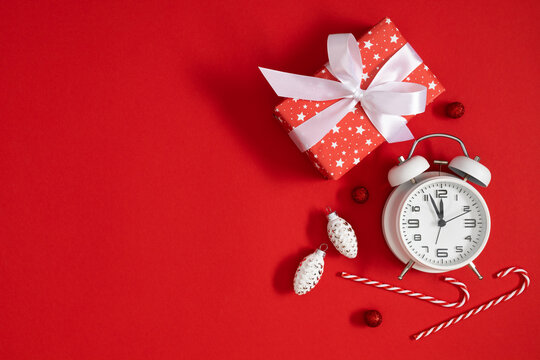Christmas flat lay with red gift box, white satin bow, vintage alarm clock, candy canes, and pinecone ornaments on vivid red background with creative negative copy space for holiday text.