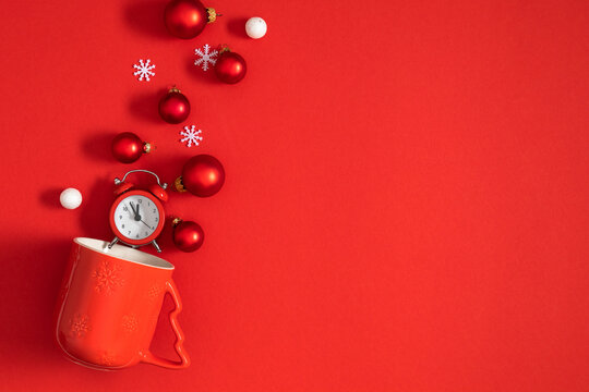 Christmas flat lay with red mug exploding with small alarm clock, bauble ornaments, and white snowflakes on vivid red background with creative negative copy space for holiday text design.