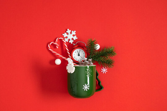 Christmas concept flat lay with green mug filled with fir branches, red alarm clock, candy canes, and snowflakes on a bright red background with dynamic negative copy space for text.