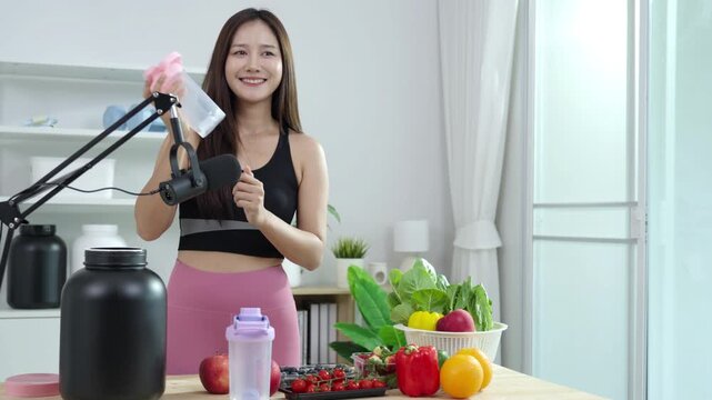Smiling Asian fitness influencer recording a health podcast, scooping pink protein powder into a shaker bottle. Woman vlogger creating sports nutrition and healthy diet content with fresh food.