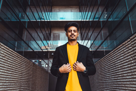 Moroccan man standing in modern urban setting, wearing a yellow t shirt and blazer, expressing confidence and fashion