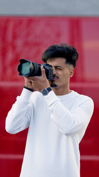 Young Moroccan man engrossed in photography, holding a professional camera with both hands, focusing intently on a red background