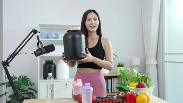 Smiling Asian fitness influencer recording a health podcast, scooping pink protein powder into a shaker bottle. Woman vlogger creating sports nutrition and healthy diet content with fresh food.