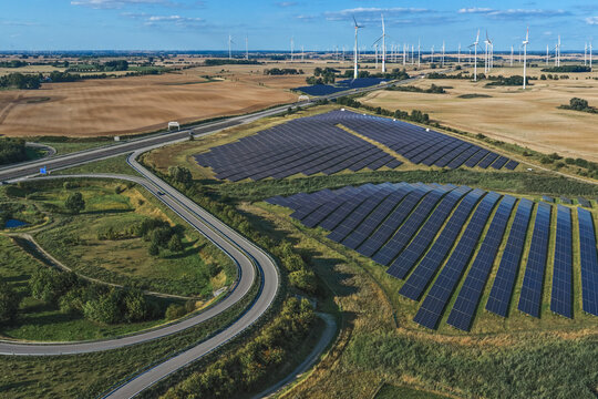 Aerial view of a large solar farm with rows of photovoltaic panels situated next to a highway, surrounded by wind turbines and agricultural fields in Uckermark, Brandenburg, Germany.