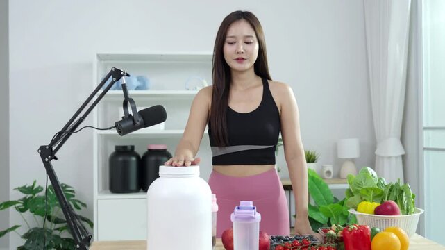 Smiling Asian fitness influencer recording a health podcast, scooping pink protein powder into a shaker bottle. Woman vlogger creating sports nutrition and healthy diet content with fresh food.