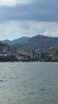 A picturesque coastal town nestles against verdant hills overlooking a calm, blue sea under a partly cloudy sky. Switzerland, Lugano, Lake Lugano