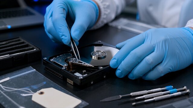 Forensic investigator examining open hard drive with tweezers. Digital data recovery and cybercrime investigation concept. Close up of hands in blue gloves