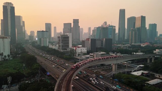 Jakarta, Indonesia - 23 July 2026: Aerial view of the city skyline with an MRT train moving along the elevated tracks above a busy highway at sunset.