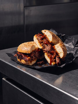 Halved Pulled Beef Brisket Bagel Sandwich with Barbecue Sauce Served on Black Paper over Industrial Stainless Steel Food Truck Kitchen Counter Top