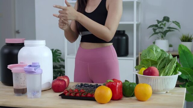 Smiling Asian fitness influencer recording a health podcast, scooping pink protein powder into a shaker bottle. Woman vlogger creating sports nutrition and healthy diet content with fresh food.