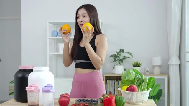 Smiling Asian fitness influencer recording a health podcast, scooping pink protein powder into a shaker bottle. Woman vlogger creating sports nutrition and healthy diet content with fresh food.