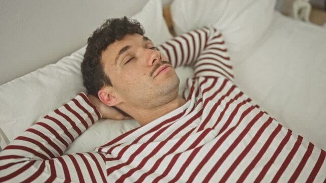 Man lying on bed with arms behind head and eyes closed, wearing striped pyjama top in studio; calm rest.