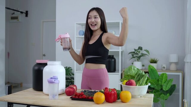 Smiling Asian fitness influencer recording a health podcast, scooping pink protein powder into a shaker bottle. Woman vlogger creating sports nutrition and healthy diet content with fresh food.