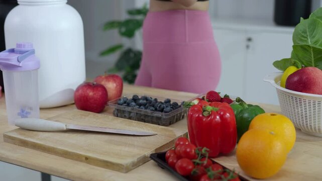 Smiling Asian fitness influencer recording a health podcast, scooping pink protein powder into a shaker bottle. Woman vlogger creating sports nutrition and healthy diet content with fresh food.