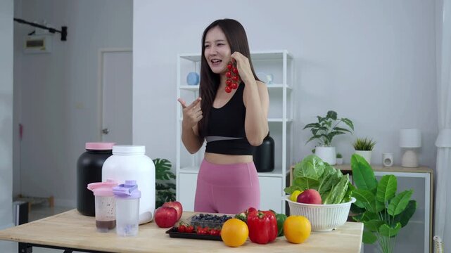 Smiling Asian fitness influencer recording a health podcast, scooping pink protein powder into a shaker bottle. Woman vlogger creating sports nutrition and healthy diet content with fresh food.