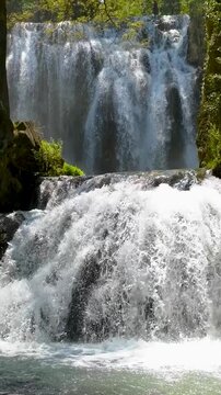 Vista vertical en plano medio de una cascada natural de dos niveles cayendo violentamente sobre rocas en un r&iacute;o rodeado de &aacute;rboles y vegetaci&oacute;n frondosa en un d&iacute;a soleado. Monasterio de Piedra.