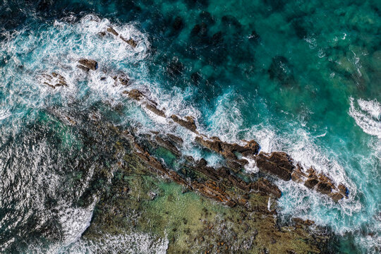 Aerial downward view of ocean waves crashing over the rugged rocky coastline at Brooms Head, capturing white surf, exposed rock formations and surrounding Pacific Ocean along the Clarence Valley 