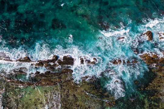 Aerial downward view of ocean waves crashing over the rugged rocky coastline at Brooms Head, capturing white surf, exposed rock formations and surrounding Pacific Ocean along the Clarence Valley coast