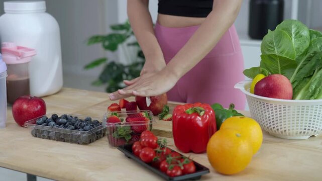 Smiling Asian fitness influencer recording a health podcast, scooping pink protein powder into a shaker bottle. Woman vlogger creating sports nutrition and healthy diet content with fresh food.