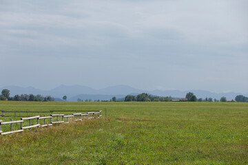 Naklejka premium Expansive green field with a wooden fence, trees, and hazy mountains under a cloudy sky.