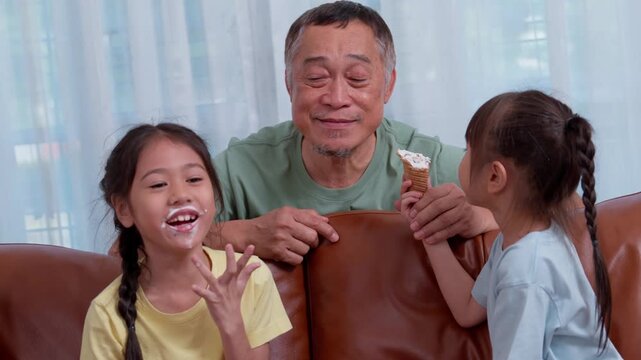 Grandfather enjoying a snack with two young granddaughters on a sofa at home. The natural family interaction conveys affection, caregiving, childhood, and multigenerational connection.
