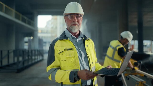 Real Estate Building Construction Site: Portrait of Male Architect Analyzing Development Data Using Laptop Computer, Looking at Camera and Smiling Crew Builds Behind. Slow Motion