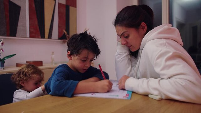 Parent tutors her son through a written exercise at the family table while his toddler sibling looks on, illustrating patient mentorship, focused study and connected home life