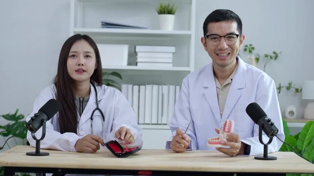 Asian male dentist holding a teeth model explaining oral care to a female doctor during a medical podcast interview. Recording a dental healthcare audio broadcast in a modern clinic studio.