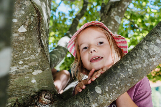 Smiling cute girl in striped hat looking through tree branches. Happy family with children on summer nature holiday.