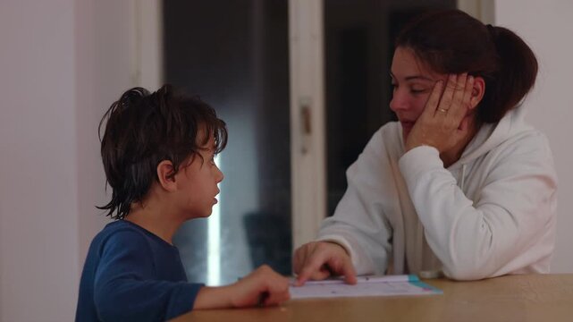Mother prompts her son to recall and repeat school material at the home table in the evening, strengthening memory, comprehension and confidence through active learning