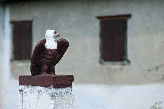 Vusanje, Gusinje Municipality, Montenegro - 28 July 2005: View of a painted eagle statue perched on a stone pillar in front of a rustic building with dark wooden window shutters.