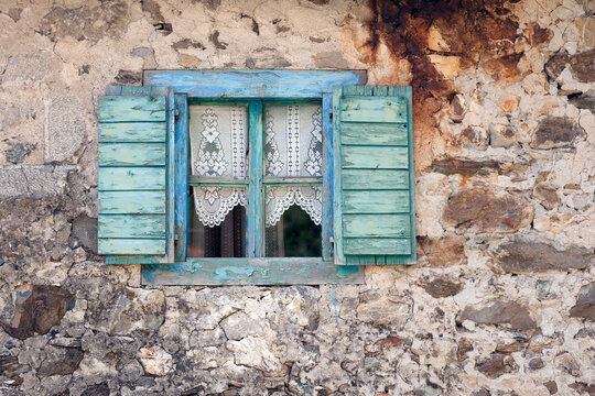 Vusanje, Gusinje Municipality, Montenegro - 28 July 2005: View of a weathered stone wall featuring a rustic window with blue wooden shutters and delicate lace curtains.