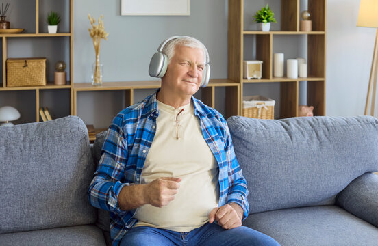Senior man with headphones on sofa enjoying music. Content retiree rests in a bright living room, smiling, moving to beat for wellness and lifestyle uses. Theme of cozy home relaxation.