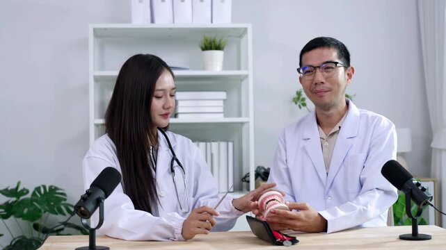 Asian male dentist holding a teeth model explaining oral care to a female doctor during a medical podcast interview. Recording a dental healthcare audio broadcast in a modern clinic studio.