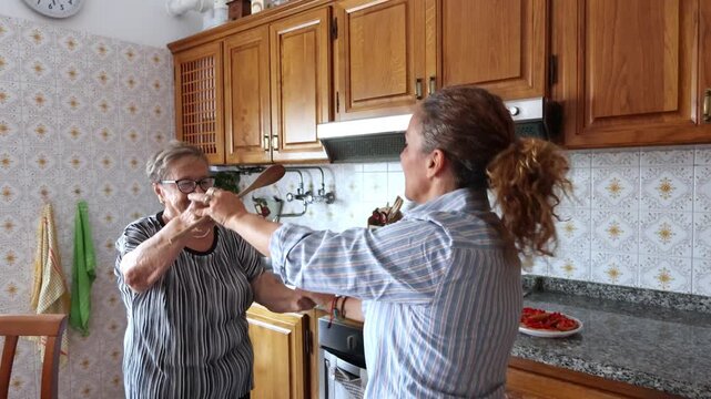 Senior mother and her adult daughter laughing and dancing together in the kitchen while preparing food, enjoying a beautiful moment of family bonding and happiness in their home