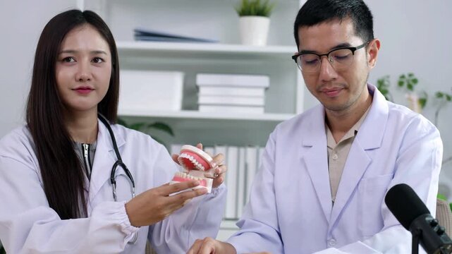 Asian male dentist holding a teeth model explaining oral care to a female doctor during a medical podcast interview. Recording a dental healthcare audio broadcast in a modern clinic studio.