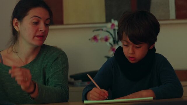 Mother explains a writing assignment to her schoolboy at the family table as he pauses with frustration, promoting patience, resilience and supported learning