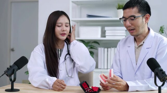 Asian male dentist holding a teeth model explaining oral care to a female doctor during a medical podcast interview. Recording a dental healthcare audio broadcast in a modern clinic studio.