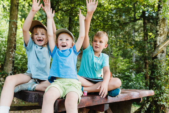 Three joyful children sitting on a picnic table raising their arms in celebration at a forest camp.