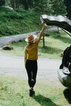 Happy young woman in a hat closing car trunk on a forest road during a joyful summer road trip adventure.