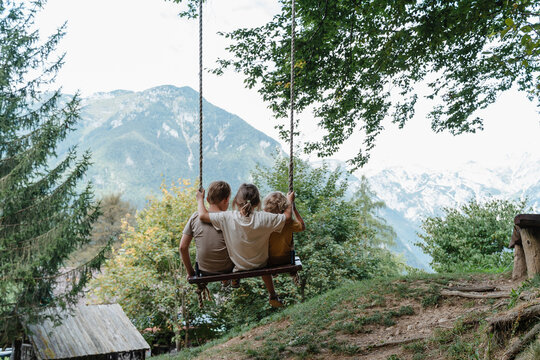 Three joyful children sitting together on a wooden tree swing overlooking a mountain valley.